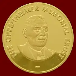 A golden medallion with an embossed image of a mustached man. Surrounding the bust in bold uppercase type is embossed the words, THE OPPENHEIMER MEMORIAL TRUST.