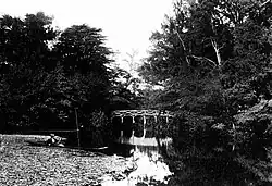 A black-and-white photograph of a body of water, with a couple in a punt (left) and a white wooden bridge (centre)