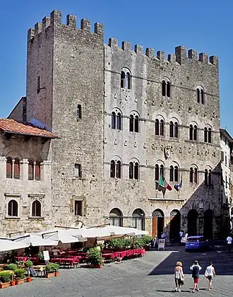 An imposing four-storey stone building with battlements and rows of paired windows, facing onto a town square.