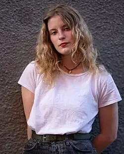 Paloma leans against a gray wall, her white shirt tucked in to her belted jeans.