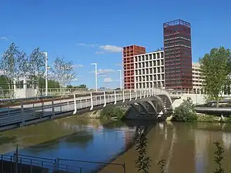 A view of a footbridge over a river. On one end of the bridge are the apartment buildings of the Olympic Village.