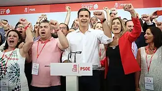 Pedro Sánchez wearing a white shirt and cheering with multiple people