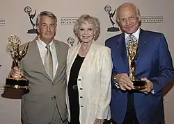 Three people standing in front of a banner are smiling at the camera. Two men are holding Emmy statuette with a woman in the middle