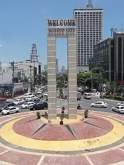 A marble monument, monolithic in structure, situated on a traffic island painted red and yellow. The monument has text near its top which says: "WELCOME QUEZON CITY"