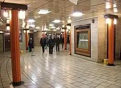 View of part of circular concourse with cream tiled floor, coffered flat ceiling and travertine stone walls. Faceted orange columns support bronze light fittings.