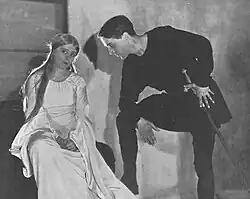 youngish white woman and man in medieval costume, she seated, he standing leaning towards her