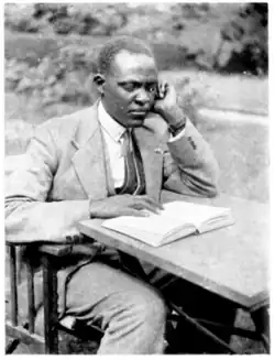 Parmenas Githendu Mockerie in a three-piece suit. He is sitting at a camp table in an English garden reading a book.