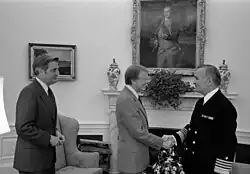 In a black and white image, Jimmy Carter and Stansfield Turner shake hands in front of the ivy, while Walter Mondale stands to the side.