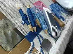 Tapestry tools on a loom: mirror, bones (wrapped with yarn), scraper (with short teeth), heavy comb (double-ended, to batten the weft), and awl (tip hidden).