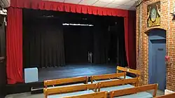 There is a black, proscenium-style stage platform surrounded by bright red curtains. In the foreground are benches with blue cushions. On the right is a brick wall with blue door.