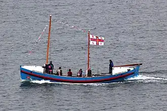 A small blue boat seen from the side. It has a mast at the front and a mast in the centre from which a white and red RNLI flag is flying.