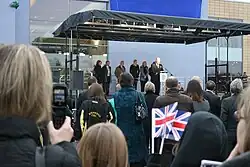 Rebecca Adlington and parents stand on a canopied, raised platform outside of the refurbished swimming pool looking on as the then-mayor of Mansfield at a microphone addresses spectators