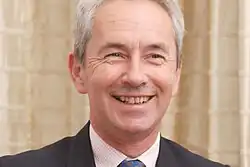 Official colour portrait photograph of Richard Stagg, smiling, standing in front of elaborate stonework
