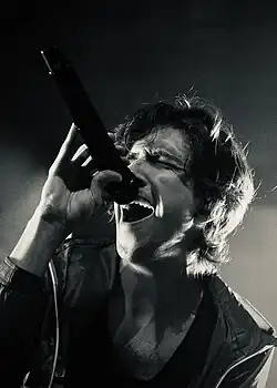 A shoulders-up portrait of Ricky Montgomery in concert. He is wearing a tank top and light raincoat, and is singing passionately into a microphone.