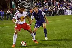 A man in a white soccer jersey defends a soccer ball from a man in a blue jersey, while a crowd of people watches.