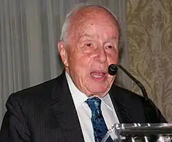 Rolf Noskwith, as an elderly man, speaking at a podium, with microphone partially obscuring his face.
