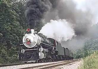 A green steam locomotive with a silver smokebox pulling a rake of passenger coaches in an area of trees