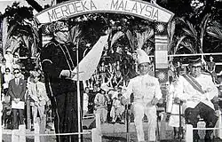 A man reading from a large piece of paper in front of an arch saying "MERDEKA MALAYSIA"