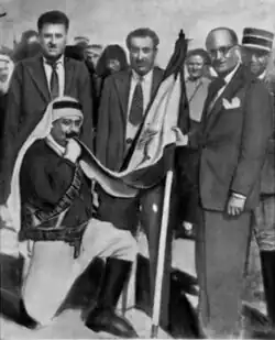 Emir Majid Arslan kneeling and kissing a flag, with Sabri Hamadeh and other officials standing nearby.