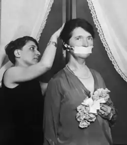 A woman stands still while another woman puts a gag over the first woman's mouth