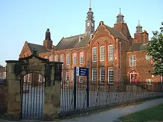 A large red-bricked building with many chimneys, with a small yard and sandstone arch in front