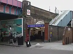 A railway on a brick viaduct crosses a road on a steel bridge, with an entrance below a blue sign reading "SHEPHERD'S BUSH MARKET STATION" in white letters