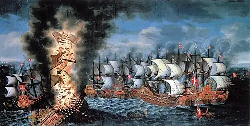 A colorful oil painting showing a large group of sailing warships engaged in battle. In the foreground to the left, a very large ship flying a Swedish flag is listing heavily and a huge explosion is shattering her structure and throwing men and equipment upwards in together with flames and black smoke