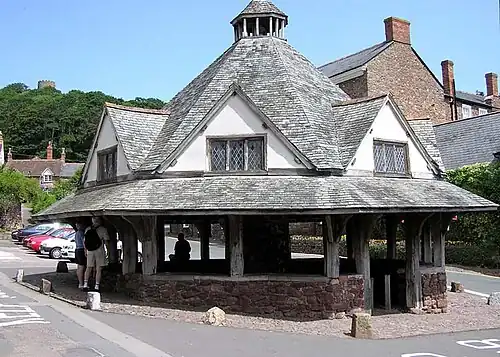 A small single-story building with a pyramid shaped roof, to the side of a road lined with buildings. Some private small cars visible. Trees in the distance with the skyline of Dunster Castle.
