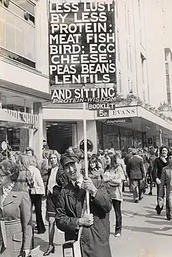 Green in Oxford Street in 1974, carrying a sign with three placards, which read, in capitals: "Less lust, by less protein: meat fish bird; egg cheese; peas beans lentils", followed by: "and sitting protein-wisdom", then, finally, hanging below the second placard: "booklet 5p".