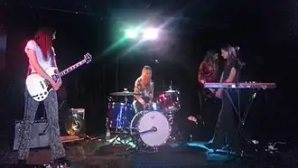 A shot of four women on stage in mid-performance. The first woman at right is cradling her guitar without playing it, she stands looking to her left: towards the second woman. She wears over shoulder-length red-brown hair, a white t-shirt with grey and black paisley-styled long pants and black shoes. The second woman is seated at her drum kit with her arms at her lap behind a drum. A microphone in a stand is above her. She wears slightly shorter hair, which is blonde-light brown, her shirt has a white background with a brown and grey design, and jeans. The third woman is partly obscured, she is playing her bass guitar and is turned to her left. She wears longer dark brown hair, a dark red-brown shirt with three-quarter sleeves over dark pants. The fourth woman is behind her keyboard and a microphone on a stand. The keyboard has writing: "nord electro 4". She stands turned to her right, facing the second woman. He arms are crossed with hands clasped at her waist. He legs are almost crossed, too. Her hair is shorter than the previous woman but has a similar colour. Her t-shirt is dark blue with jeans and dark shoes. Surrounding the women is stage equipment including speakers, instruments, electrical cords and a pair of stage lights (in the centre above the second woman).