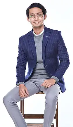 Sufian Suhaimi wearing grey pants, white shirt, grey sweater, and a blue striped blazer, sitting on a tall stool or chair, grinning directly at camera