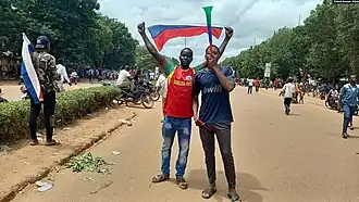Two Burkinabe men show their support for Russia during the reopening of the Russian embassy in Ouagadougou in 2023 in the middle of a street, waving a Russian flag above them with other supporters in the distance.