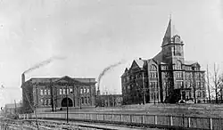 Two buildings stand side by side on a hill. The one on the left is two stories, with two smokestacks behind it. The one on the right is the larger, taller Tech Tower building.