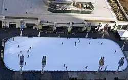 An ice skating rink with a few skaters and a large metallic sculpture in the background