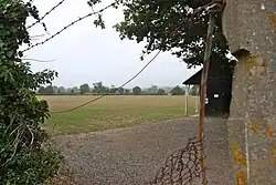 Through a wired fence, there is a grass playing field and a cricket pavilion to the right.