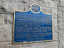 A blue plaque on a stone wall. The plaque has a yellow border, and is mostly rectangular in shape, with the long end oriented horizontally. However, the top side has a camel hump in the centre, with a circle centred at the top of the hump. Inside the circle is an Ontario coat-of-arms. The plaque reads: THE MACDONALD CARTIER FREEWAY This plaque commemorates the completion of the Macdonald-Cartier Freeway (Highway 401), the longest freeway operated without tolls by a single highway authority in North America. Covering 510 miles between Windsor on the Canada–US border and the Ontario-Quebec boundary, it serves the richest economic region in Canada. In January 1965, it was named by The Honourable John Robarts, Premier of Ontario, in honour of the two founding architects of the Confederation of Canada, Sir John A. Macdonald and Sir George-Étienne Cartier. This site is located on the last section of construction, consisting of 15 miles between Ivy Lea and Highway 2, which was completed on October 11, 1968.