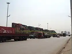 line of heavy trucks going north, background a container yard
