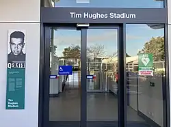 a photograph of an information panel about Hughes and a set of sliding doors with the words "Tim Hughes Stadium" above them
