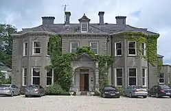 It is a large grey building with big windows. There are trees growing around the front door. Five cars are parked on both sides of the front door.