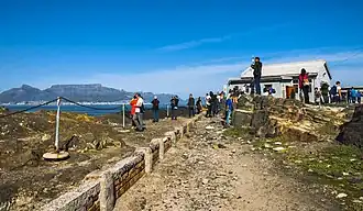A straight stone pathway through a rocky area, elevated in places from the walkway, with a low retaining wall and chain fence on the left. Beyond is an area of ocean; at the far background on the left is a large flat rocky mountain with a peaked one at its left. Closer to the camera is a small building on the right; people are milling around it and the paths, some taking pictures