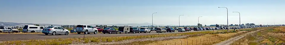 A road with vehicles of various types stopped on it, all facing right, stretching out over a grassy, flat landscape away from the viewer, getting farther away to the left. In the foreground is a dirt road and fence