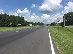 A four-lane asphalt road in a wooded area with some farmland, with a sign that says North 113