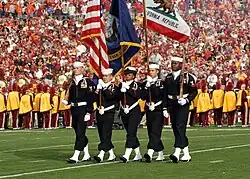 Photograph of five members of the Naval Base Ventura County Honor Guard parading the colors, the University of Southern California Marching Band is in the background wearing yellow and performing the national anthem during the 2008 Rose Bowl game