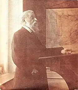 An elderly bearded man in a suit standing stoically at a lectern