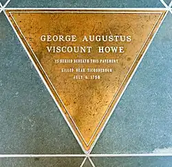 A golden triangular marker set amidst triangular green stone tiles. Engraved on it is "George Augustus Viscount Howe" in large type, and in smaller type below "Is buried beneath this pavement ... Killed near Ticonderoga July 6, 1758"