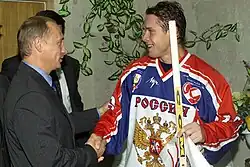 An ice hockey player in his early thirties shakes hands with a middle-aged man dressed in a dark suit. The hockey player wears a white, red and blue jersey labeled "РОССИЯ" and holds a hockey stick.