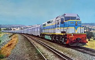 In a rural scene, a two-tone blue diesel locomotive with yellow striping and red headstock at the head of the Indian Pacific train comprising stainless steel passenger cars