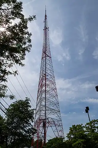 A three-sided steel lattice tower, painted orange and white, rising high above trees and power lines
