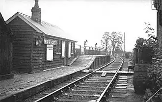 A small wooden hut labelled "Westcott". In front of the hut is a deserted low wooden railway platform with a short section at a much greater height; the only objects on the platform are three large lamps. A single railway track leads past the platform; the line branches immediately past the end of the platform. A cat is asleep on the railway track.