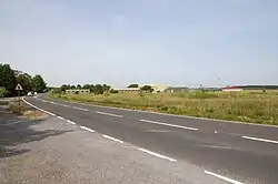 A photo of the airfield, in the foreground is a roadway, with a lay-by on the left hand side. To the right of the image is grass and vegetation, and in the centre of the image off in the distance are a number of buildings.